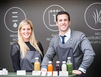 a female and a man standing behind a row of cold pressed juices